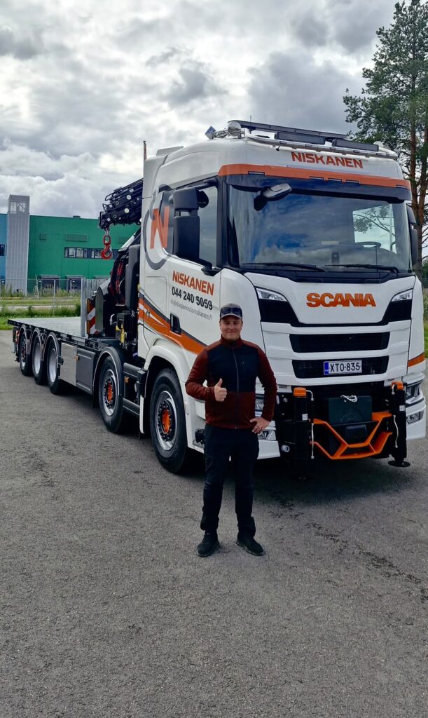 Man posing in front of a white Scania heavy truck with orange Niskanen branding and a flatbed trailer behind him.