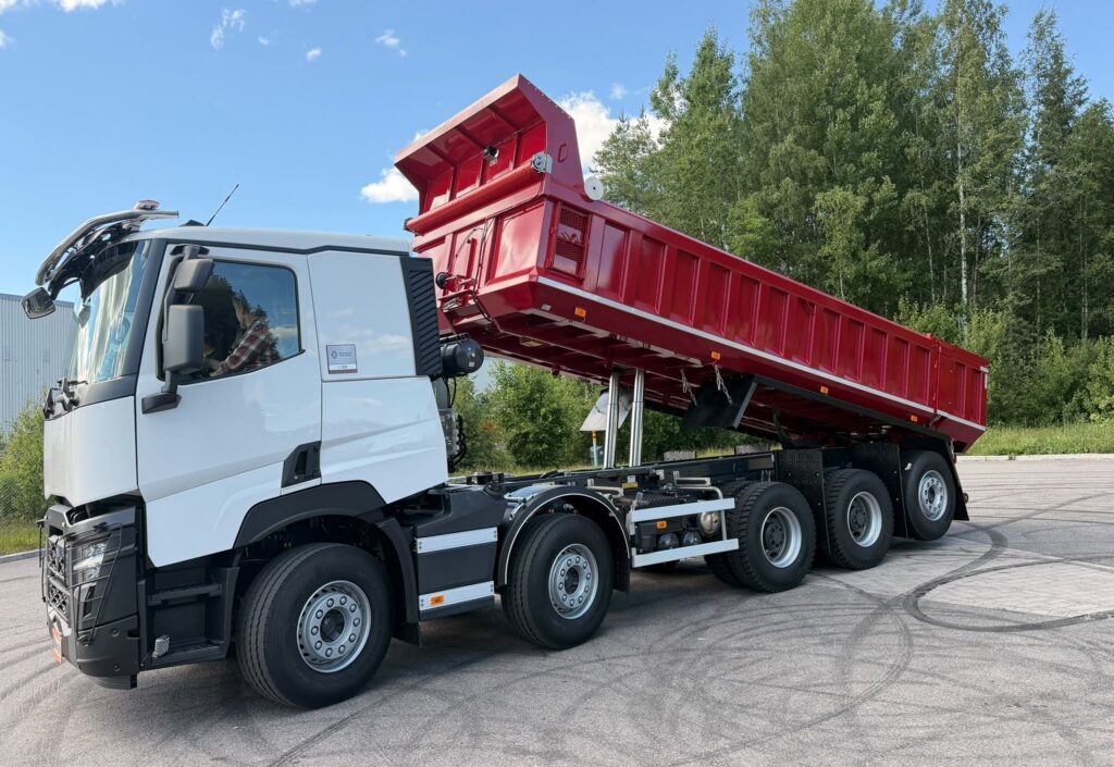 White heavy-duty semi truck with a red tipper/dump trailer raised in the air, parked on a paved lot with green trees in the background, under a blue sky.