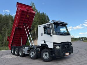 White multi-axle dump truck with its red dump bed raised for dumping, parked on a paved road with trees in the background.