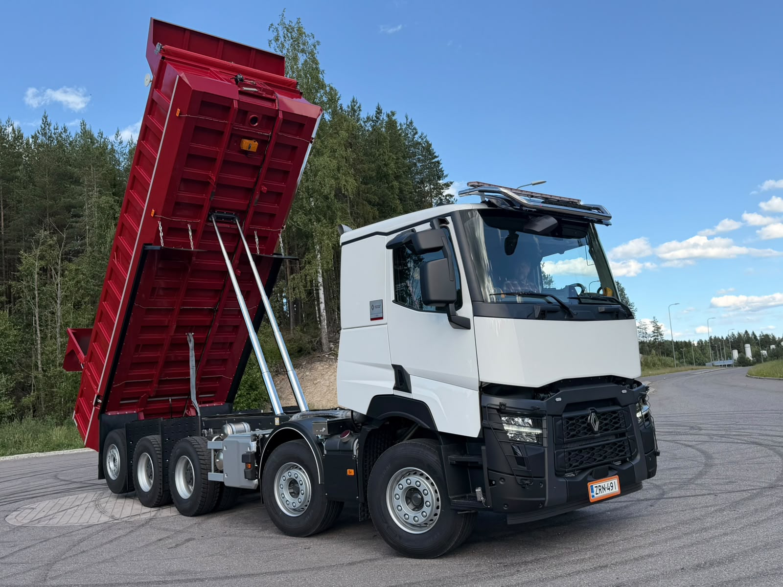 White multi-axle dump truck with its red dump bed raised for dumping, parked on a paved road with trees in the background.