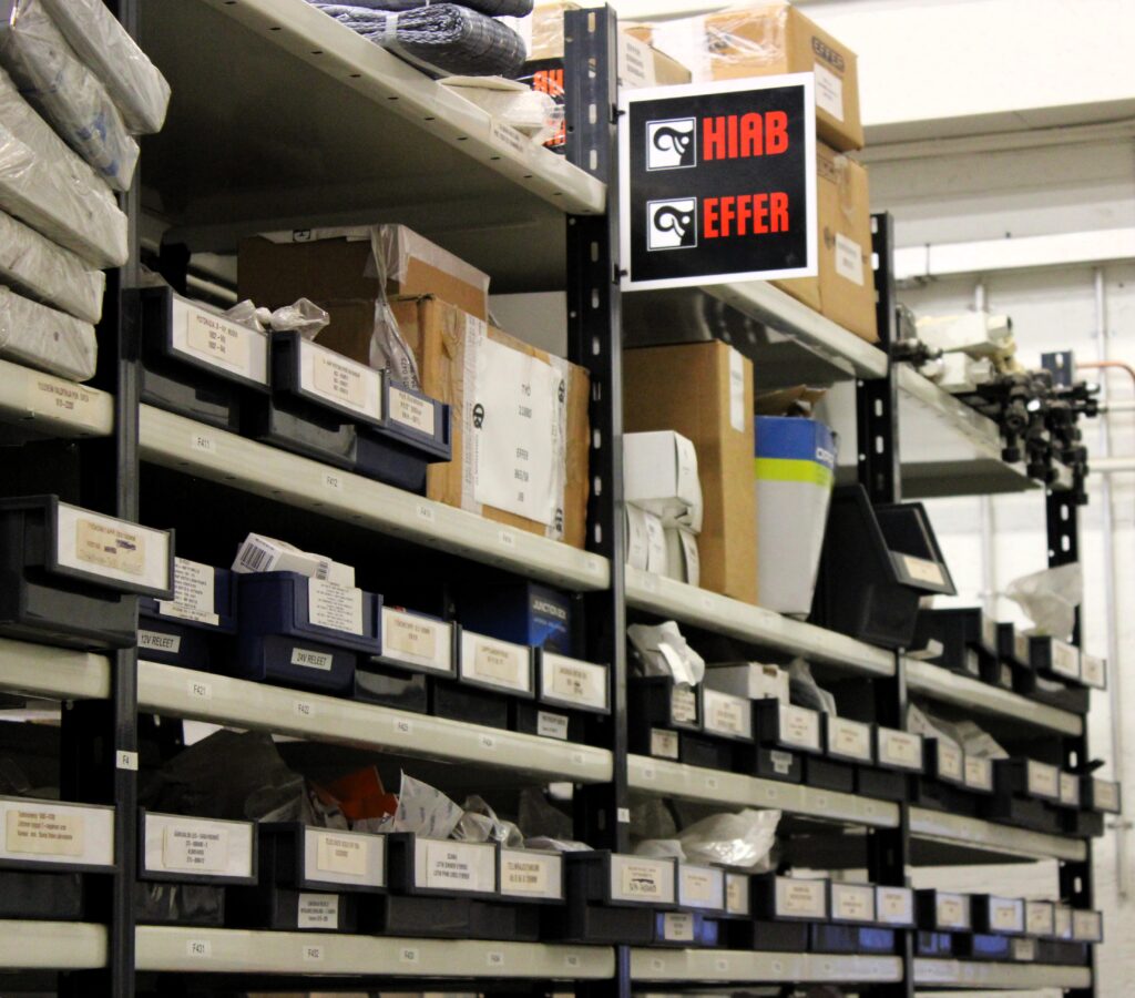 Warehouse shelving with labeled boxes; a black HIAB EFFER sign hangs on the rack.